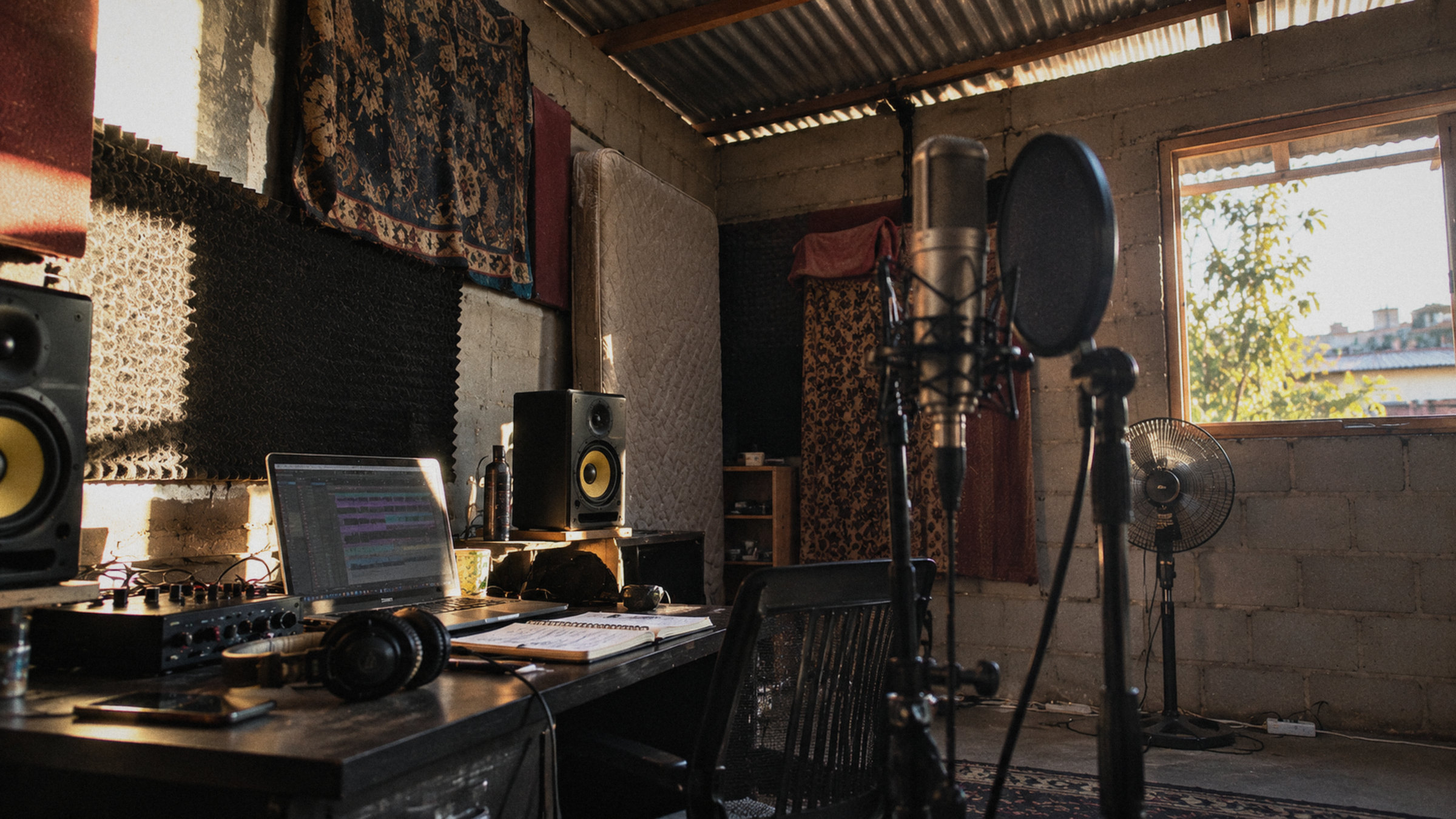 A small makeshift recording studio in a concrete-block room with a corrugated metal roof: a condenser microphone with a pop filter, studio monitors, a laptop and audio interface on a desk, a mattress and fabric on the walls for sound dampening, and a standing fan by a sunlit window.